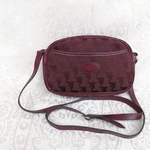 Puede incluir: Un bolso de hombro burdeos con una correa larga. El bolso presenta un patrón repetitivo y un pequeño bolsillo frontal. La correa es ajustable y tiene una hebilla dorada. El bolso tiene un parche de cuero con el nombre de la marca.