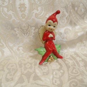 May include: A ceramic figurine of a red elf with a pointed hat, sitting on a green holly leaf base with three gold-coloured berries. The elf has painted facial features, including dark eyes and eyebrows, and a pair of small, pale wings on its back.