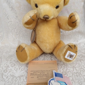 Könnte beinhalten: Ein goldfarbener Teddybär mit brauner Nase und dunklen Augen. Der Bär hat ein Etikett und einen kleinen Aufnäher am Fuß. Ein Reisepass und ein Aufkleber mit einer amerikanischen Flagge sind ebenfalls sichtbar.