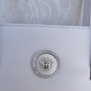 Puede incluir: Un bolso blanco con dos asas y un emblema decorativo. El emblema es un c&iacute;rculo plateado con un dise&ntilde;o de cabeza de Medusa en el centro, rodeado de cristales brillantes. El bolso est&aacute; hecho de un material liso y claro.