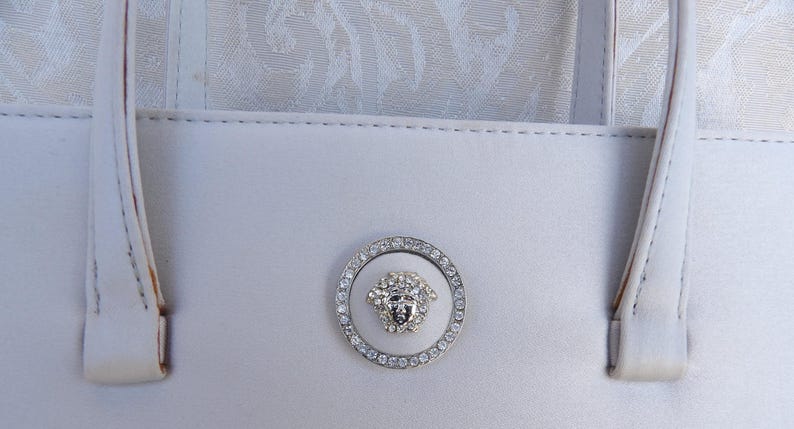 Puede incluir: Un bolso blanco con dos asas y un emblema decorativo. El emblema es un c&iacute;rculo plateado con un dise&ntilde;o de cabeza de Medusa en el centro, rodeado de cristales brillantes. El bolso est&aacute; hecho de un material liso y claro.