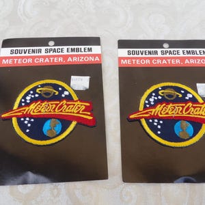 Könnte beinhalten: Zwei runde gestickte Aufnäher auf schwarzen Präsentationskarten. Jede Karte trägt die Aufschrift SOUVENIR SPACE EMBLEM METEOR CRATER, ARIZONA. Die Aufnäher zeigen einen dunkelblauen Hintergrund, einen gelben Rand, einen Planeten und Sterne.