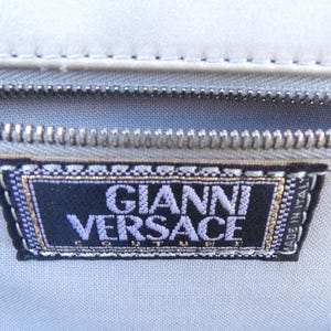 Puede incluir: Primer plano de una tela gris claro con una cremallera plateada y una etiqueta negra. La etiqueta dice "GIANNI VERSACE" en letras moradas con un borde dorado. Tambi&eacute;n se pueden ver las palabras "MADE IN ITALY".