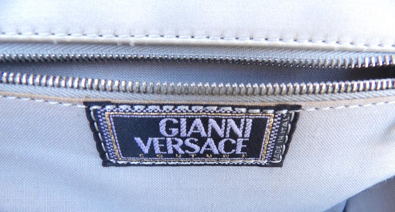 Puede incluir: Primer plano de una tela gris claro con una cremallera plateada y una etiqueta negra. La etiqueta dice "GIANNI VERSACE" en letras moradas con un borde dorado. Tambi&eacute;n se pueden ver las palabras "MADE IN ITALY".