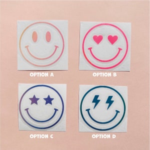 Pu&ograve; includere: Quattro adesivi quadrati con disegni di faccine sorridenti. L'opzione A ha una sfumatura di rosa e arancione. L'opzione B ha occhi a cuore rosa. L'opzione C ha occhi a stella. L'opzione D ha occhi a fulmine. Ogni adesivo ha la lettera dell'opzione sotto.
