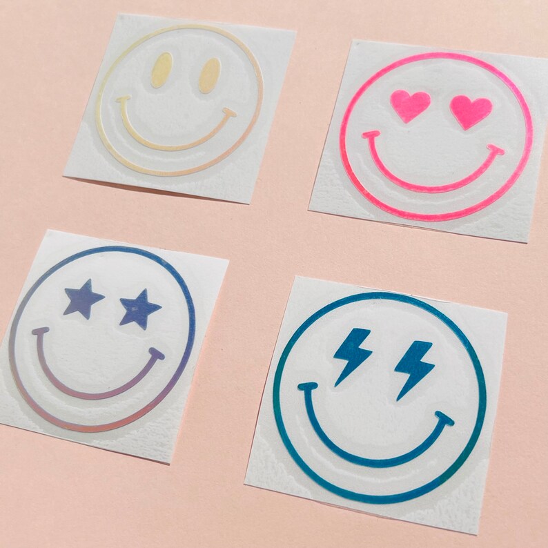 Pu&ograve; includere: Quattro adesivi a forma di faccina sorridente con diversi disegni. Uno ha gli occhi gialli, un altro gli occhi a cuore rosa, un terzo gli occhi a stella e l'ultimo gli occhi a fulmine. Ogni adesivo &egrave; su sfondo bianco.