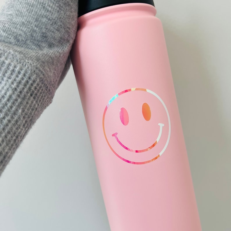 Pu&ograve; includere: Una borraccia rosa chiaro con un coperchio nero e un adesivo a forma di faccina sorridente colorata. La faccina sorridente ha un contorno bianco e un motivo rosa, arancione e giallo all'interno.