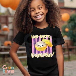 Könnte beinhalten: Schwarzes T-Shirt mit einer Grafik eines lächelnden Knödels in einer gelben Schüssel, mit dem Text "Hey there Dumplin'" in Weiß und Gelb. Das Design enthält rosa und lila Streifen. Das T-Shirt wird von einem lächelnden Kind getragen.