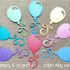 May include: A collection of nine die-cut paper balloons in various colors including light green, teal, blue, yellow, pink, purple, and red. Each balloon features a textured surface, a small cutout near the top, and a swirling, curly string design.