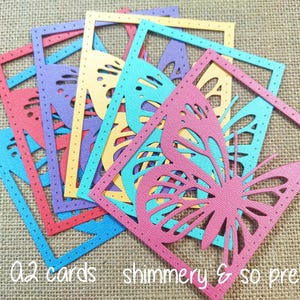 May include: A set of six layered die-cut cards featuring a butterfly design in shades of blue, pink, yellow, and purple. The cards are designed for A2 cards and have a shimmery finish.