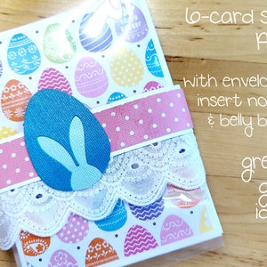 May include: A set of six Easter cards with colorful egg designs, packaged with envelopes, insert notes, and a belly band. The cards are decorated with a white lace trim and a pink polka dot ribbon. The set includes a blue bunny cutout.