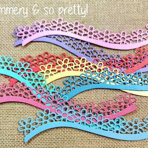 May include: A collection of die-cut paper borders in various colors including pink, purple, red, yellow, and blue, featuring a repeating daisy flower pattern along a wavy edge. The text "shimmery & so pretty!" is printed in white at the top.
