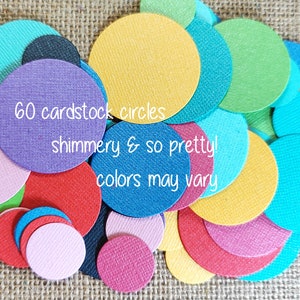 May include: A collection of 60 assorted cardstock circles in various vibrant colors including purple, yellow, red, blue, green, and pink. Each circle features a textured surface and a subtle shimmer. The text 60 cardstock circles, shimmery & so pretty!, colors may vary is printed across the center of the overlapping paper shapes.