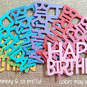May include: A collection of die-cut cardstock cake toppers featuring the text Happy Birthday in various colors including yellow, green, teal, blue, purple, red, and light pink. Each piece has a textured surface finish and a cutout letter design.