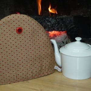 Peut inclure: Une théière en céramique blanche avec un couvercle et une anse est posée à côté d'un chauffe-théière. Le chauffe-théière est beige avec un motif de cœurs rouges et un bouton rouge. Un foyer avec des flammes est en arrière-plan.