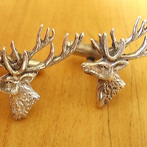 May include: A pair of silver cufflinks featuring detailed stag head designs. The stag heads are facing forward with antlers extending out to the sides.