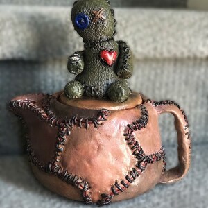Könnte beinhalten: Eine handgefertigte, skurrile Teekanne mit einem genähten Design und einer kleinen Voodoo-Puppenfigur obenauf. Die Teekanne hat eine Mischung aus Braun- und Rosatönen, mit schwarzen Nähten und einem roten Herz auf der Brust der Puppe.