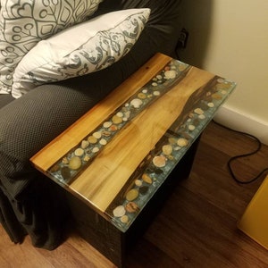 May include: A wooden side table with a clear epoxy resin top. The resin contains embedded seashells and stones. The table is next to a gray couch with pillows.