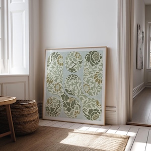 May include: A framed floral print featuring a repeating pattern of stylized flowers and foliage in shades of green, tan, and cream. The artwork is displayed in a light-colored wooden frame and leans against a white wall.
