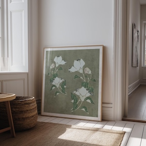 May include: A framed print displaying a botanical illustration of white flowers and green leaves on a muted green backdrop. The artwork is in a light wooden frame, leaning against a white wall in a room with natural light, creating a serene atmosphere.