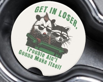 May include: Two white ceramic car coasters with a green trash can and two raccoons wearing sunglasses. The text on the coasters reads "Get in Loser, Trouble Ain't Gonna Make Itself."