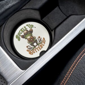 May include: A white round car coaster with a green frog wearing a cowboy hat and holding a gun. The text on the coaster says "Buckle Up, Buttercup".