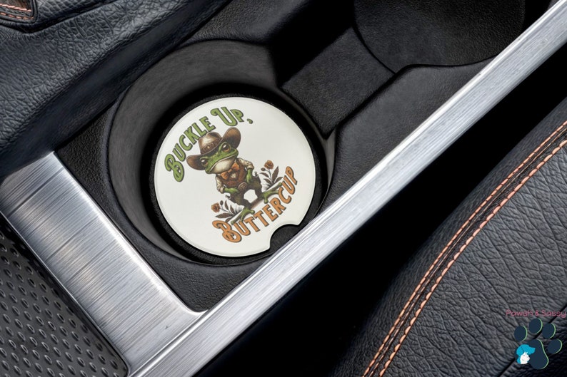 May include: A white round car coaster with a green frog wearing a cowboy hat and holding a gun. The text on the coaster says "Buckle Up, Buttercup".