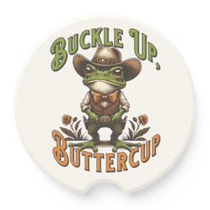 May include: A white ceramic car coaster with a green frog wearing a cowboy hat and a bow tie. The frog is holding two pistols and the text "Buckle Up, Buttercup" is written in green and orange letters.