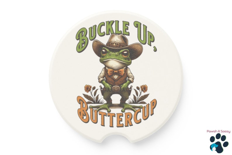 May include: A white ceramic car coaster with a green frog wearing a cowboy hat and a bow tie. The frog is holding two pistols and the text "Buckle Up, Buttercup" is written in green and orange letters.