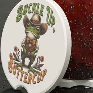 May include: White round coaster with a green frog wearing a cowboy hat and a bow tie. The frog is holding two pistols and the text "Buckle Up, Buttercup" is printed on the coaster.