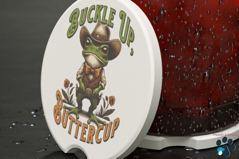 May include: White round coaster with a green frog wearing a cowboy hat and a bow tie. The frog is holding two pistols and the text "Buckle Up, Buttercup" is printed on the coaster.