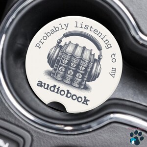 May include: A round, off-white car coaster with a black and white illustration of headphones over a stack of books. The text reads "probably listening to my audiobook."