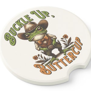 May include: A white ceramic coaster with a green frog wearing a cowboy hat and a bow tie. The frog is holding a gun and the text "Buckle Up, Buttercup" is printed on the coaster.