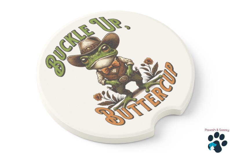 May include: A white ceramic coaster with a green frog wearing a cowboy hat and a bow tie. The frog is holding a gun and the text "Buckle Up, Buttercup" is printed on the coaster.