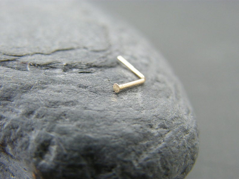 May include: A gold nose ring with a small, round bead at the end. The ring is bent at a 90-degree angle.