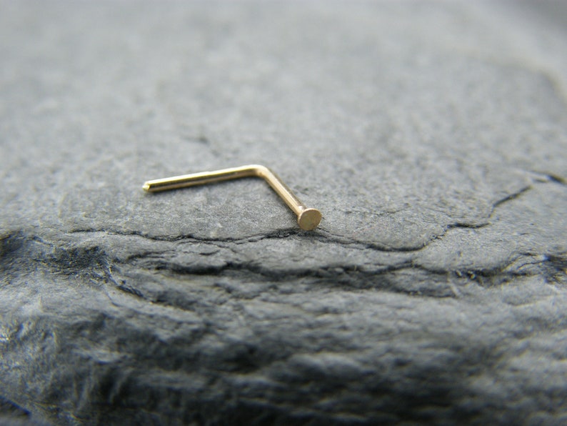 May include: A gold nose ring with a small, round ball at the end. The ring is bent at a 90-degree angle.