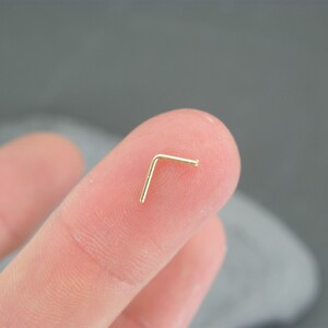 May include: A gold nose ring with a L-shaped design. The ring is held in a person's hand.