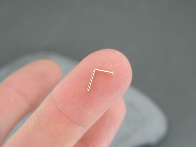 May include: A gold nose ring with a L-shaped design. The ring is held in a person's hand.