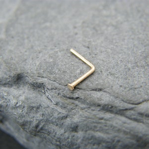 May include: A gold nose ring with a L-shaped design. The ring is made of a thin, smooth metal and has a small, rounded end.