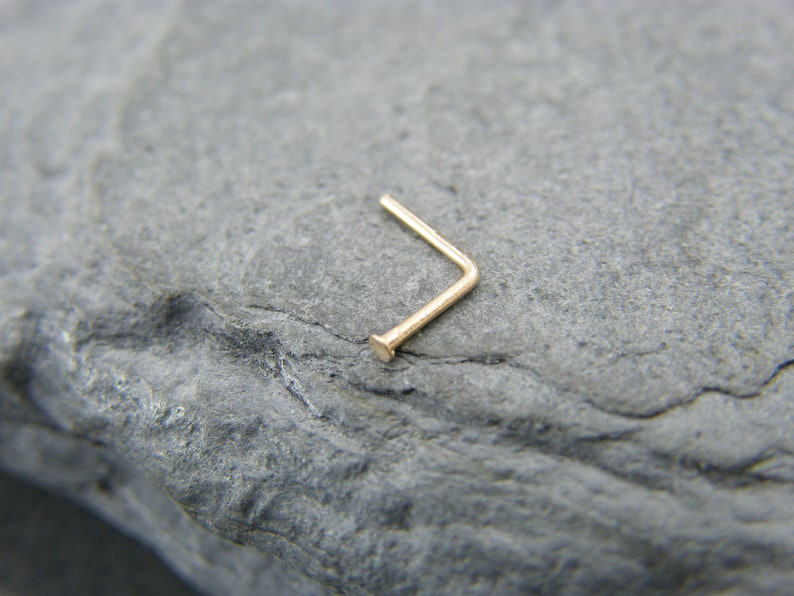 May include: A gold nose ring with a L-shaped design. The ring is made of a thin, smooth metal and has a small, rounded end.