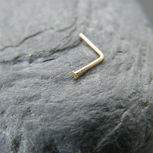 May include: A gold nose ring with a small, rounded end. The ring is bent at a 90-degree angle.