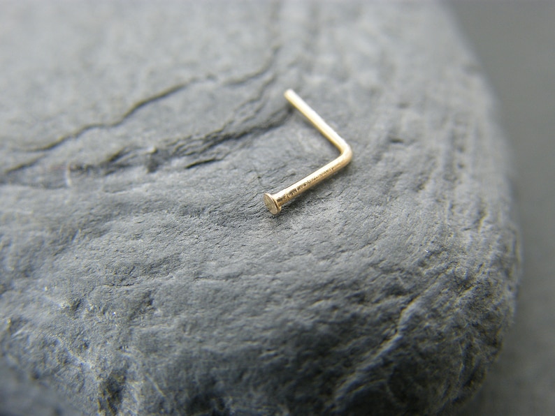May include: A gold nose ring with a small, rounded end. The ring is bent at a 90-degree angle.