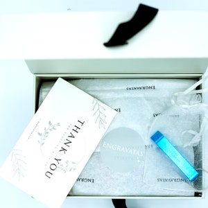 May include: A white gift box with a blue ribbon. The box is open and contains a white card with the text "Thank You for Shopping with us" and a blue keychain with the text "Engravata's" on it.