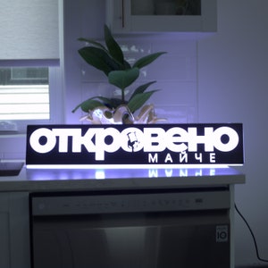 May include: A black and white sign with the text "откровено майче" and the website address "www.нe" in white letters. The sign is illuminated with white LED lights.
