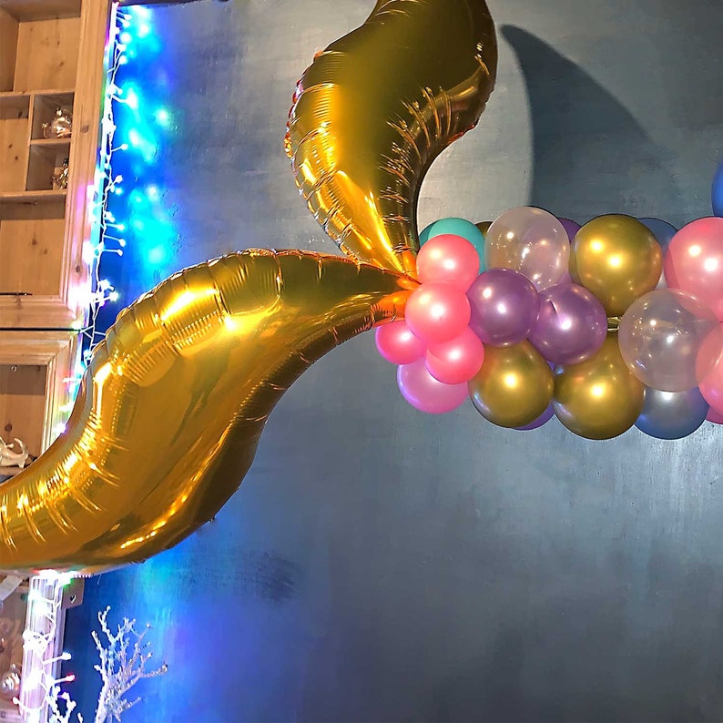 Pu&ograve; includere: Un palloncino a forma di coda di sirena in lamina dorata con un gruppo di palloncini color pastello rosa, viola, oro e turchese.