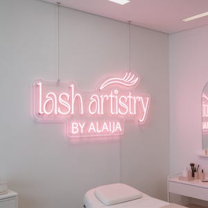 Può includere: Un'insegna al neon rosa montata su una parete con il testo "lash artistry BY ALAIJA" in una combinazione di caratteri corsivi e sans-serif, con una grafica stilizzata di ciglia sopra il testo, sospesa da due sottili cavi metallici.