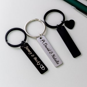May include: Three black and silver keychains with engraved names and a heart or champagne glass design. The keychains are on a white surface.