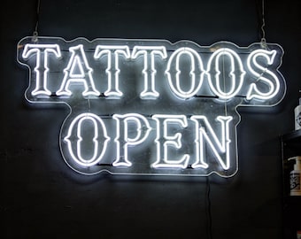 May include: A neon sign with the words "TATTOOS OPEN" in white, stylized lettering. The sign is mounted on a dark wall, and the neon lights are illuminated, creating a bright and eye-catching display. The sign is likely advertising a tattoo parlor.