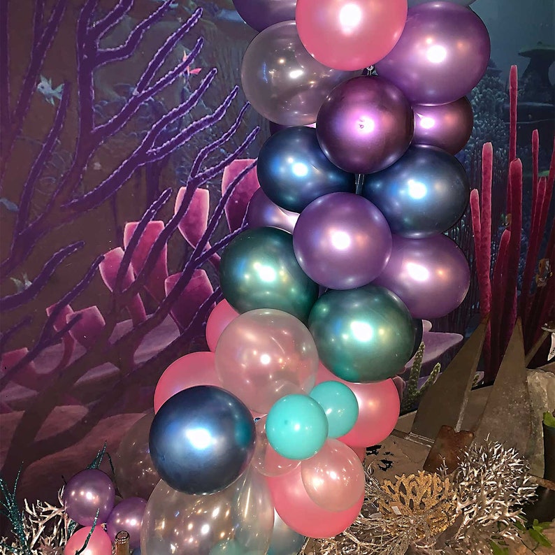 Pu&ograve; includere: Un arco di palloncini a tema sirenetta con palloncini iridescenti viola, rosa e turchese. I palloncini sono disposti a cascata, creando un'atmosfera fantasiosa e festosa.