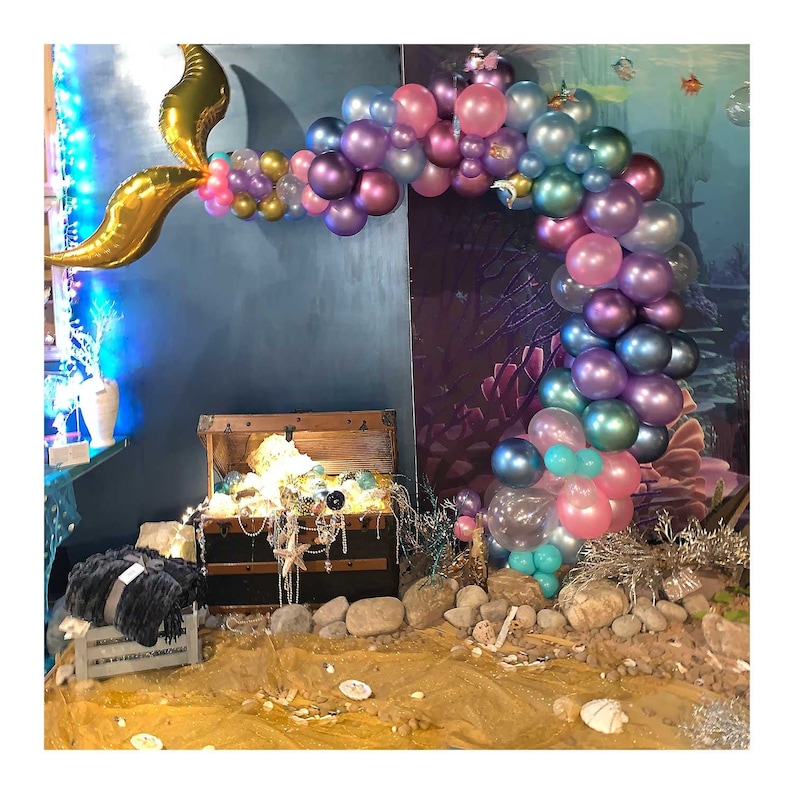 Pu&ograve; includere: Un arco di palloncini a forma di coda di sirena dorata con palloncini iridescenti viola, rosa e blu. Un forziere di legno con decorazioni dorate e bianche si trova davanti all'arco.
